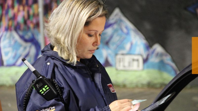 Agente loira de cabelos médios de lado conferindo a CNH ao lado de um veículo