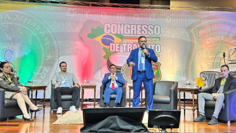 Fábio Santos falando ao microfone no palco do evento enquanto é observado pelos demais debatedores, que estão sentados
