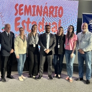 Oito pessoas em pé posando para foto em frente ao telão com a marca do Seminário Estadual de Diretores de Ensino