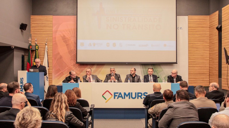 Foto horizontal colorida mostra mesa de evento com autoridades sentadas e a plateia sentada de costas