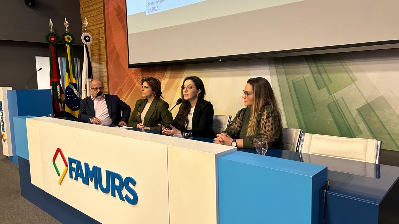 Foto horizontal colorida mostra três mulheres e um homem sentado em mesa de evento com o logo da Famurs
