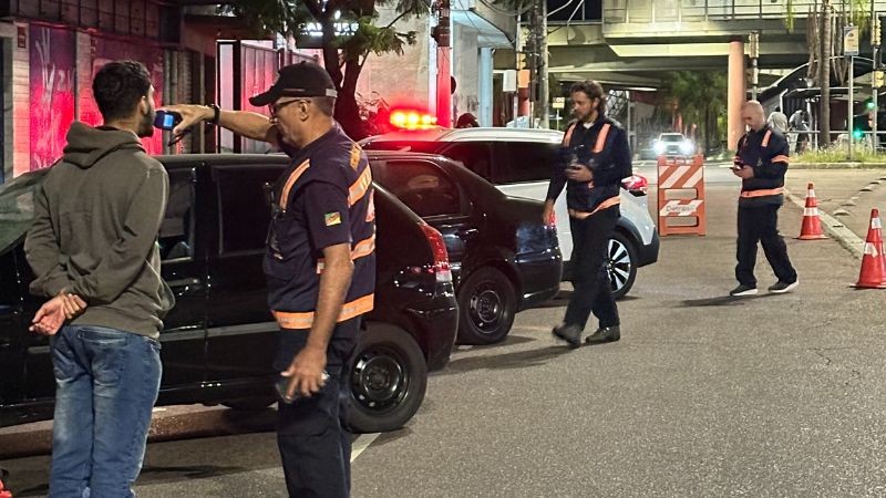 Agente da balada aplicando o teste do etilômetro em homem que está de costas para a imagem. Veículos estacionados e mais dois agentes indo em direção para fazer a abordagem.