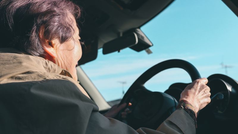 Foto horizontal colorida mostra perfil de uma pessoa idosa ao volante de um carro