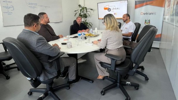 Mesa oval de reuniões com cinco pessoas em volta e uma tv ao fundo onde está escrito peça legal. Ao lado, ao fundo, banner com logo do DetranRS.