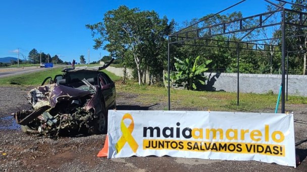 Foto de uma estrada e no recuo um veículo completamente destruído e em frente uma placa onde está escrito Maio Amarelo, junto salvamos vidas!
