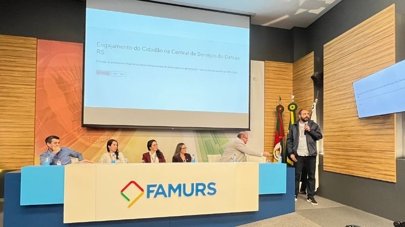 mesa do audit&oacute;rio da Famurs com cinco servidores do DetranRS sentados e um de p&eacute; apresentando sua fala. No tel&atilde;o atr&aacute;s est&aacute; escrito Engajamento do cidad&atilde;o na Central de Servi&ccedil;os do Detran.