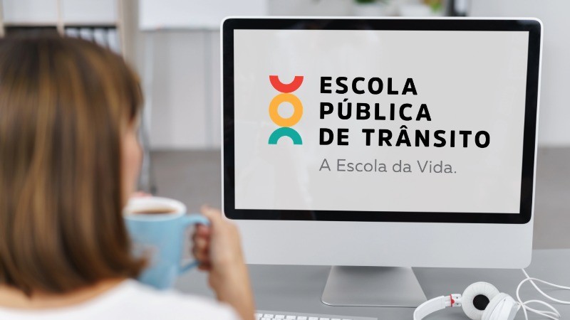 Foto horizontal colorida mostra mulher de costas com uma caneca de caf&eacute; olhando para a tela onde est&aacute; o logo da Escola P&uacute;blica de Tr&atilde;nsito do DetranRS