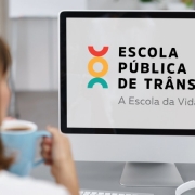 Foto horizontal colorida mostra mulher de costas com uma caneca de caf&eacute; olhando para a tela onde est&aacute; o logo da Escola P&uacute;blica de Tr&atilde;nsito do DetranRS